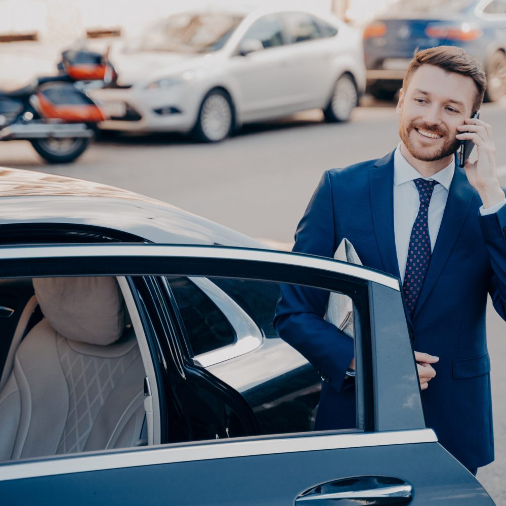 Confident smiling executive dressed in blue formal suit leaving black limousine after arriving at his workplace, talking on phone, holding newspaper while standing alone next to car with open door