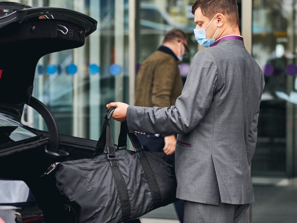 Chauffeur in a disposable face mask taking his client bag out of the car trunk