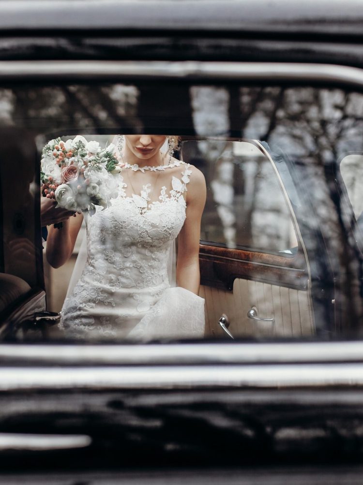 stylish wedding couple sitting in beautiful black car, creative view from window. elegant groom and bride walking and holding hands in city street, wedding day concept